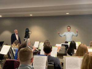 A man standing on a conductor's podium in front of people holding musical instruments. A man stands off to the side.