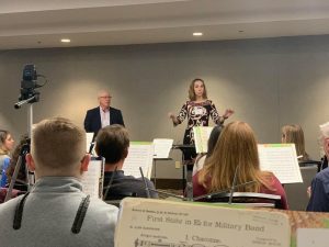 A woman standing on a director's podium holding a baton in front of people playing musical instruments. A man stands beside her.