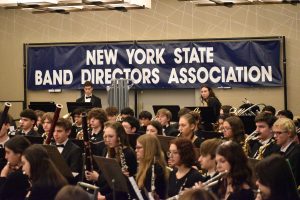 Students sitting holding musical instruments, In the background there is a sign that reads, "New York State Band Directors Association."