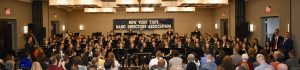 Students sitting and holding musical instruments, In the background there is a sign that reads, "New York State Band Directors Association."