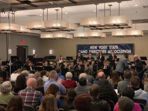 Two men shaking hands in front of a group of students holding musical instruments. In the background there is a sign that reads, "New York State Band Directors Association."