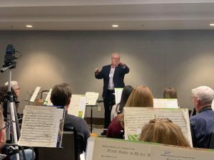 Man at a directors podium holding a baton in front of a group of people playing instruments