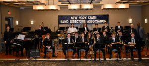 Students sitting holding musical instruments, In the background there is a sign that reads, "New York State Band Directors Association."