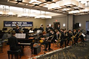 Students sitting and playing musical instruments, In the background there is a sign that reads, "New York State Band Directors Association."