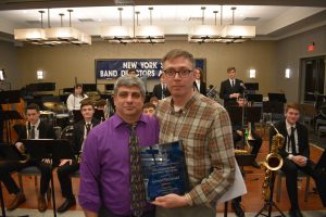 One man holding a plaque next to another man with his arm around him, In the background there is a sign that reads, "New York State Band Directors Association."