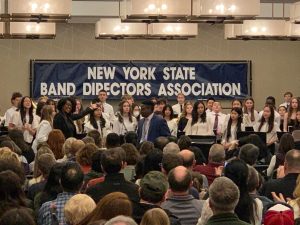 Woman standing in front of a group of students holing instruments. In the background there is a sign that reads, "New York State Band DIrectors Association."