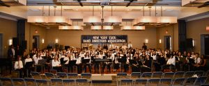 Students sitting holding musical instruments, In the background there is a sign that reads, "New York State Band Directors Association."