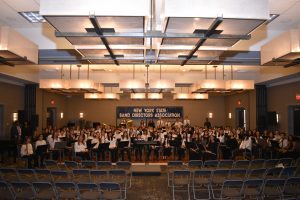 Students sitting holding musical instruments, In the background there is a sign that reads, "New York State Band Directors Association."