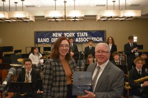 A man holding a plaque standing next to a woman. Student in the background holding musical instruments.